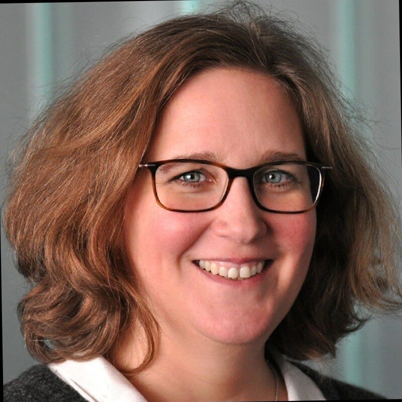 Eine Frau mit schulterlangen welligen Haaren und eckiger Brille lächelt in die Kamera.