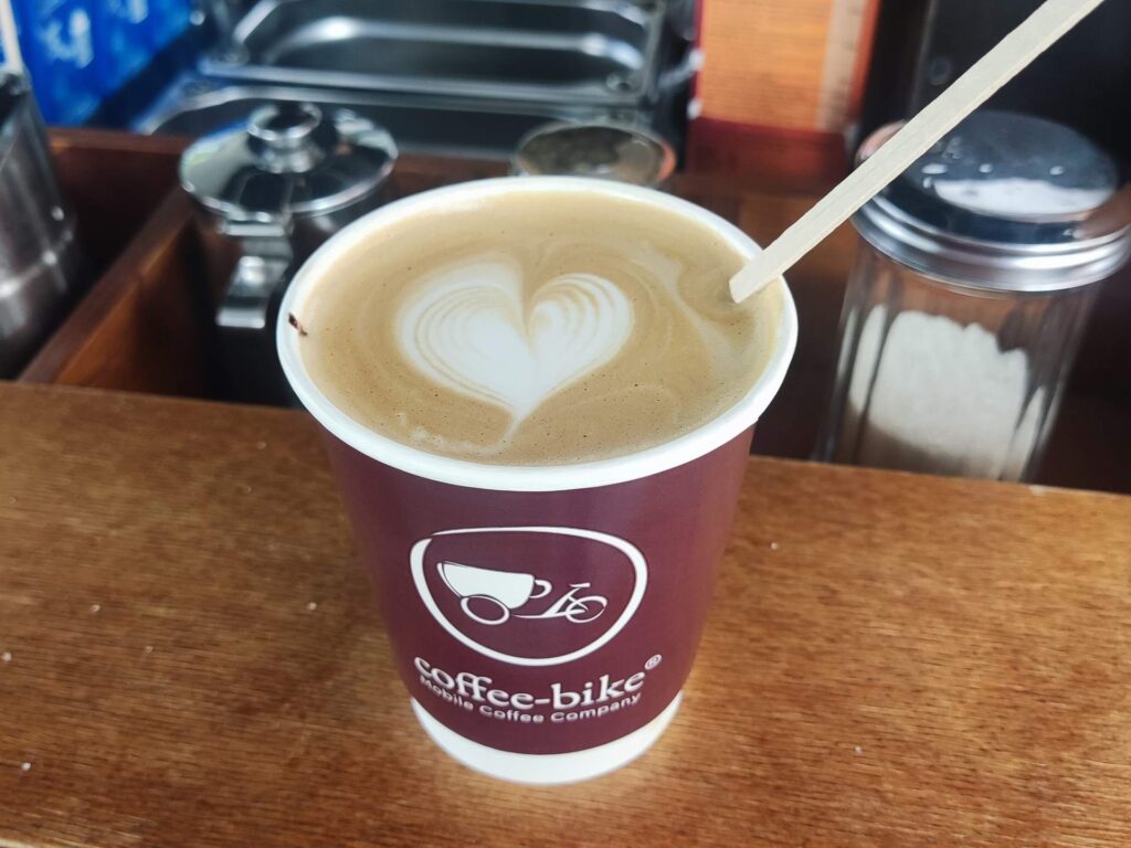 Close-up of a disposable coffee cup with a wooden stirrer. The barista has drawn a little heart with milk foam.