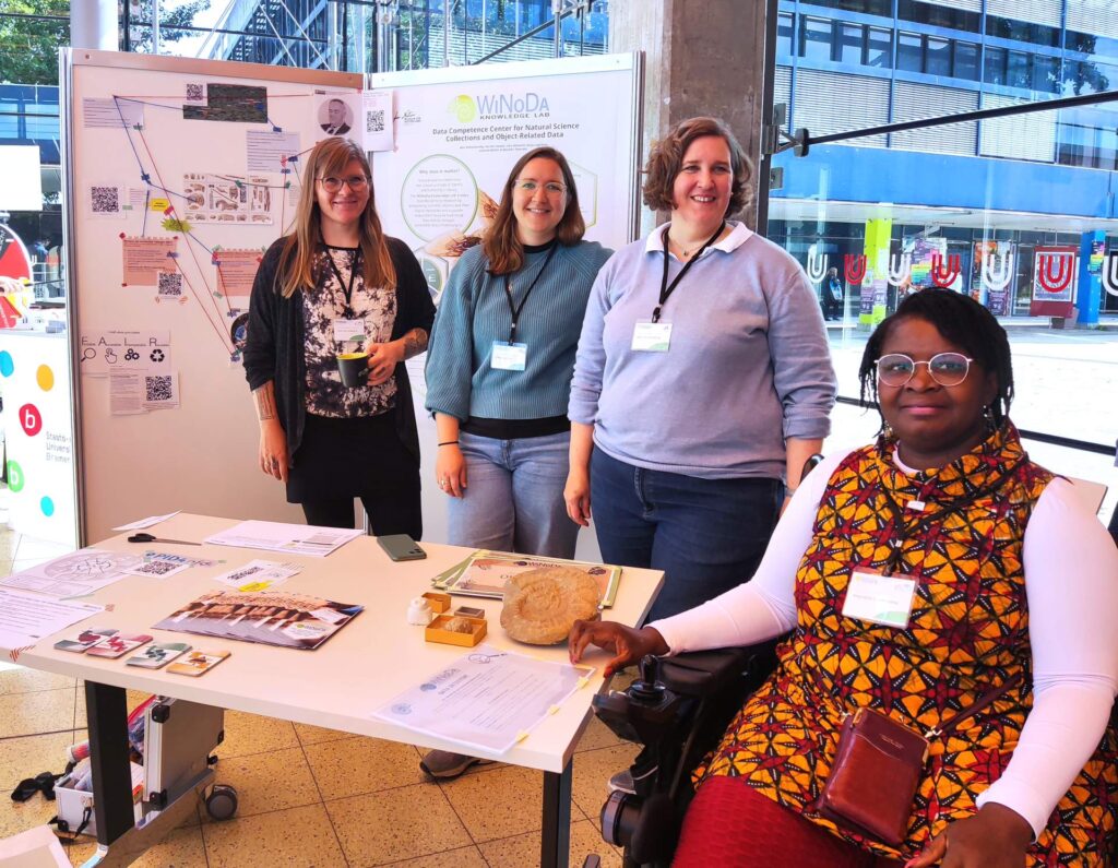 Four women behind a table with exhibits look into the camera with friendly smiles. In the background is a metaplan wall with a poster and various pieces of paper. Three of the women are standing, one is sitting in a wheelchair.