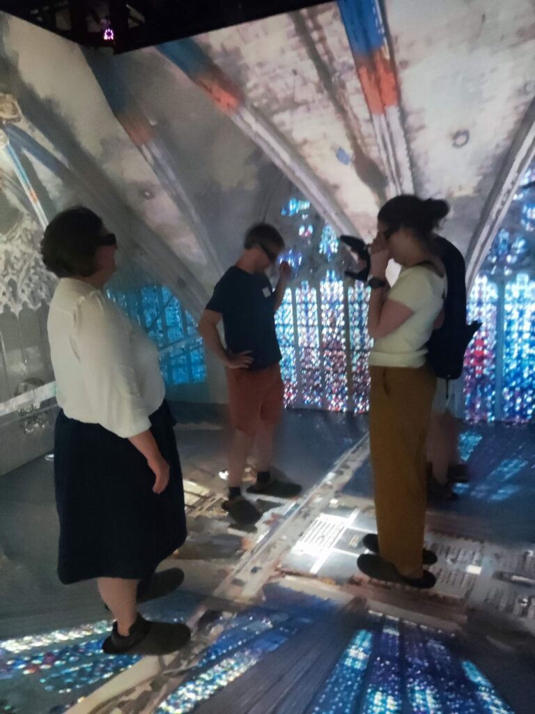 Drei Personen (plus eine verdeckte) mit VR-Brillen stehen in einem Würfel aus Bildschirmen. Es sieht so aus als schwebten sie unter der Decke des Aachener Doms.