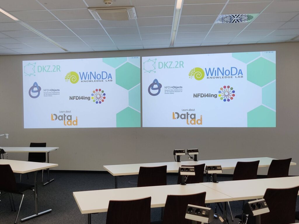 Blick in den noch leeren Veranstaltungsraum. An der Wand sieht man die Folie mit den Logos der beteiligten Organisationen.