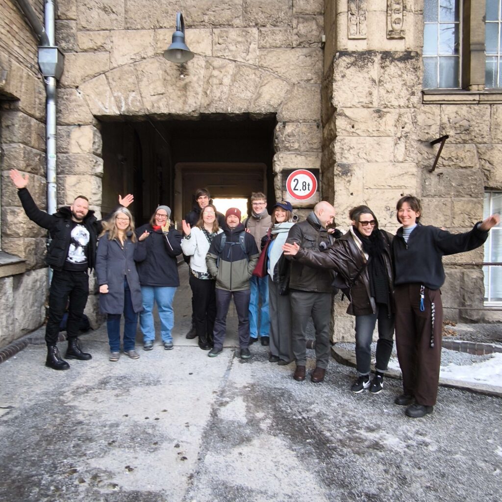 Eine Gruppe lächelnder Menschen steht vor einem Eingang und winkt in die Kamera.